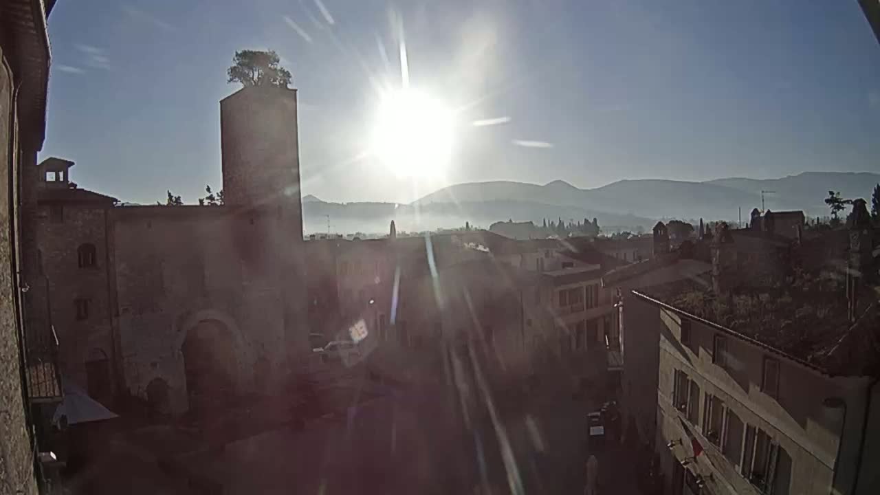 A sunlit town square is overlooked by a stone tower with a tree on top, all set against a hazy mountain backdrop.