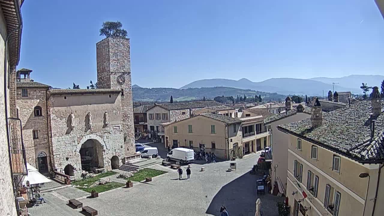 A sunny day reveals a town square in a quaint Italian village, surrounded by stone buildings and overlooked by a clock tower topped with a tree, with mountains visible in the background.