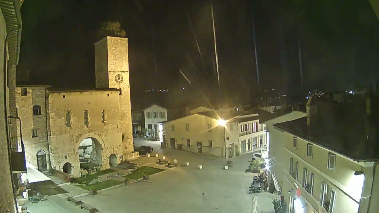 A nighttime view of a town square with a tall clock tower, illuminated buildings, and light rain falling.