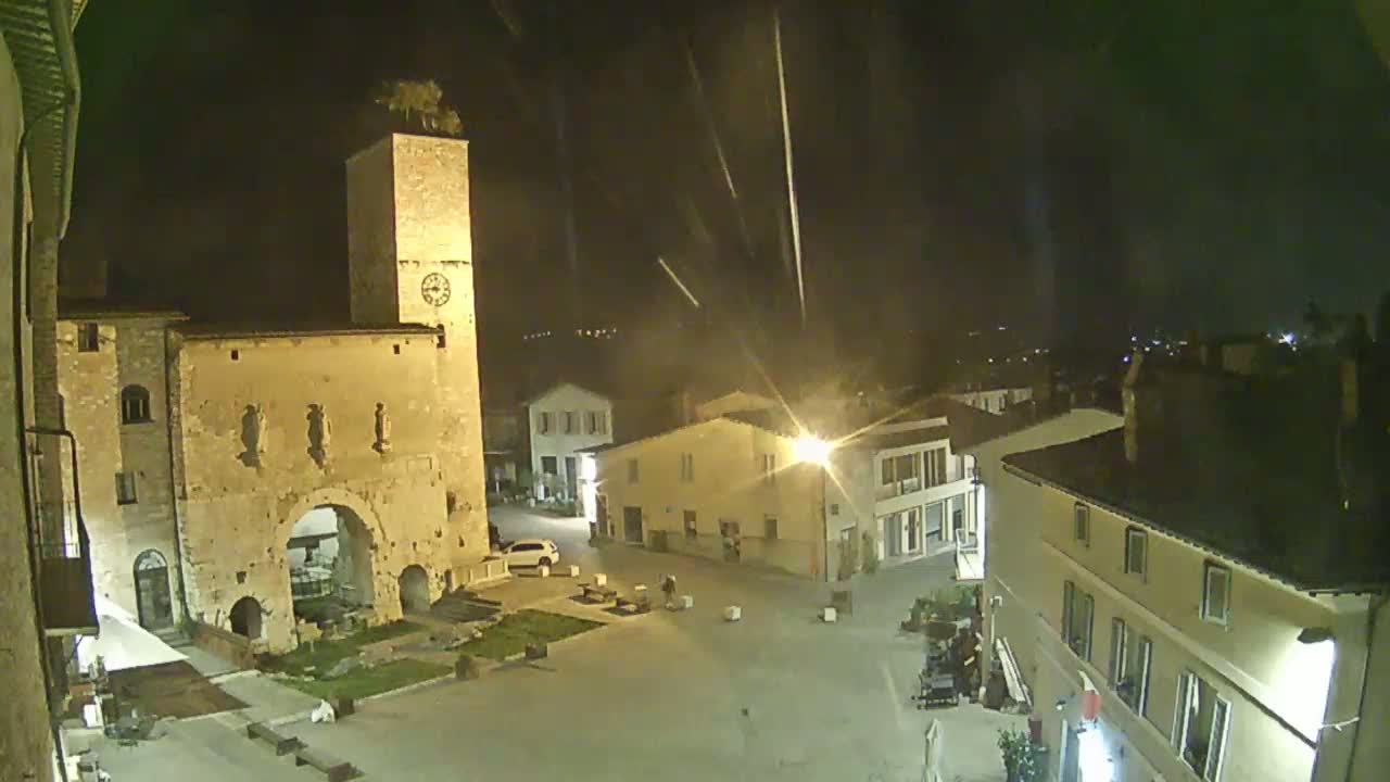 A nighttime view of a town square with an old stone building and tower, under a light rain or drizzle.