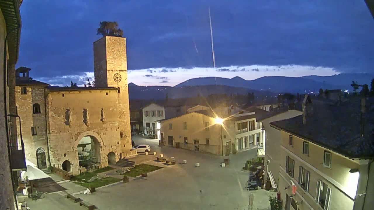 A town square at twilight, under a mostly clear, dark blue sky, is surrounded by old stone buildings, one featuring a tall clock tower with a tree on top, and overlooks a range of distant mountains.