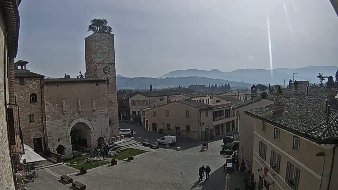A town square with stone buildings, a clock tower with a tree on top, and distant hazy mountains under a sunny sky.