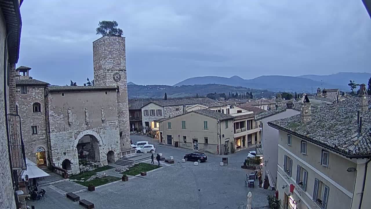 A town square in a historic Italian village, under a twilight sky, is surrounded by stone buildings, including a tall tower with a clock, and overlooks distant hills.