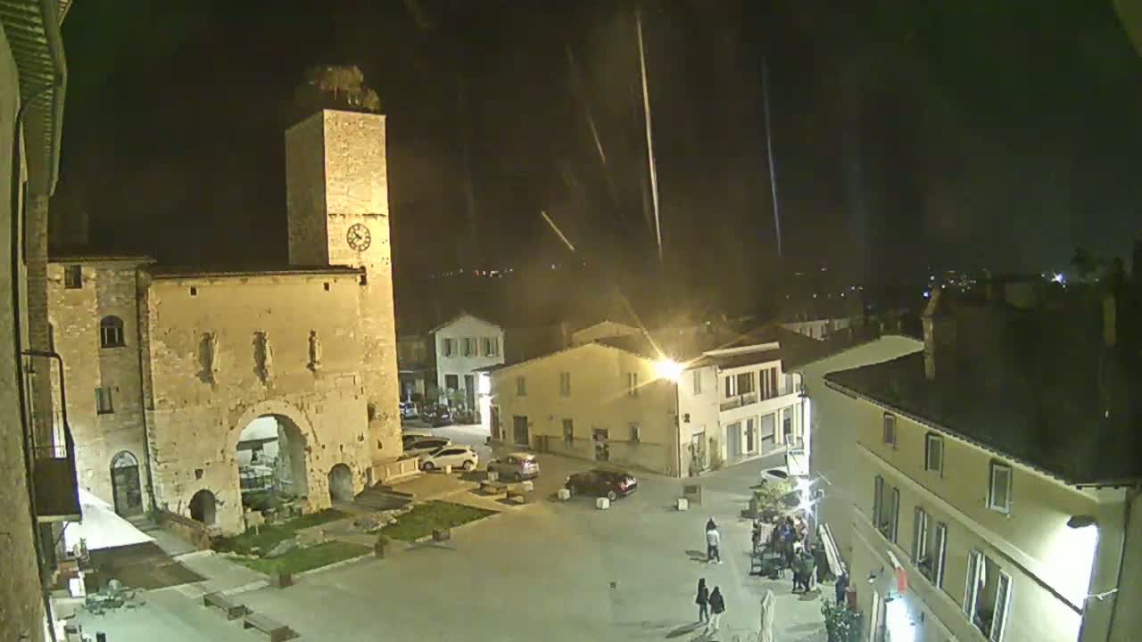 A nighttime view of a town square with several buildings, including a tall tower and a stone archway, under a light rain.