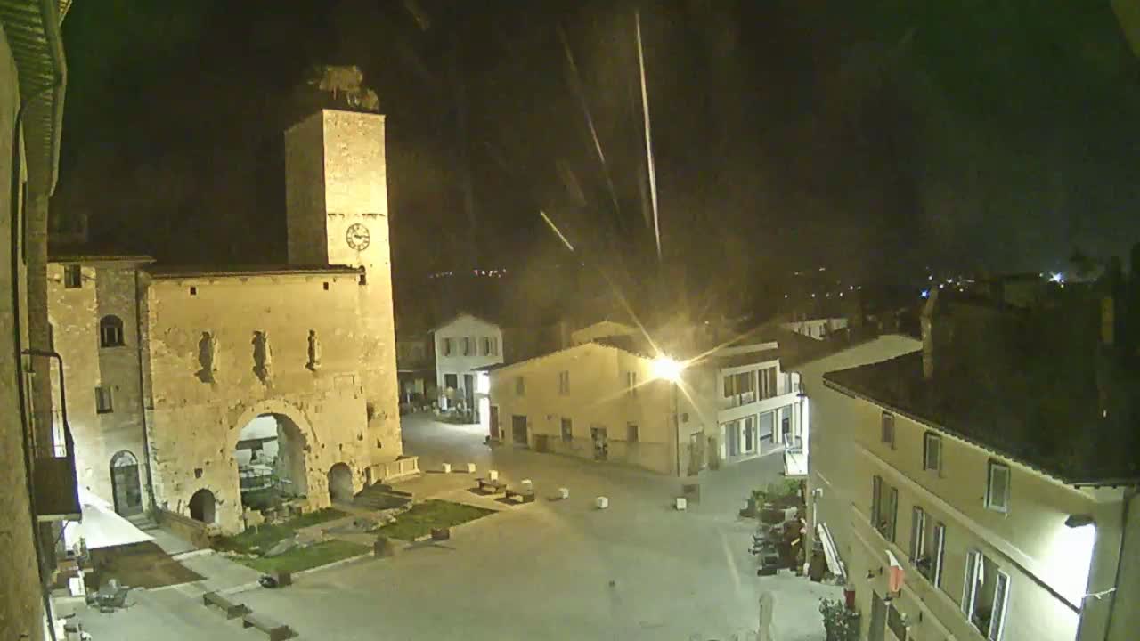 A nighttime view of a town square with a tall clock tower, several buildings, and a sparsely populated plaza under a clear, dark sky.