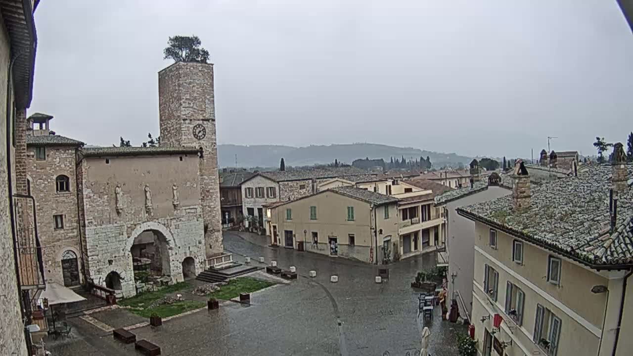 A wet town square with stone buildings, a clock tower topped with a tree, and a view of distant hills under an overcast sky.