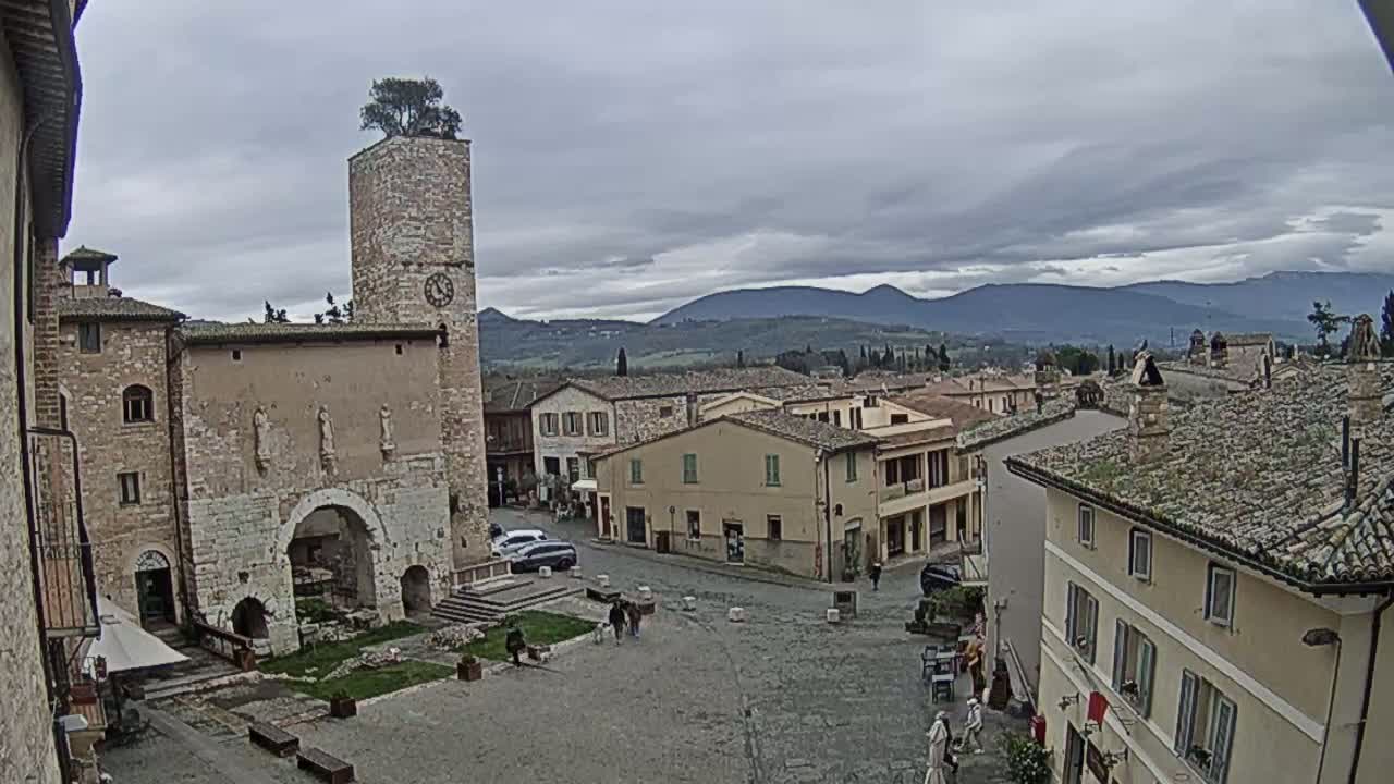 A town square in a historic Italian village, under a cloudy sky, is surrounded by stone buildings, including a tall tower with a tree on top, and overlooks a distant mountain range.