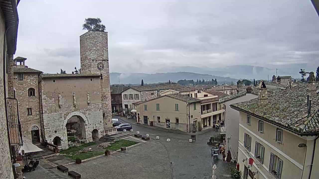 A town square with a tall clock tower, stone buildings, and a few cars, under an overcast sky.