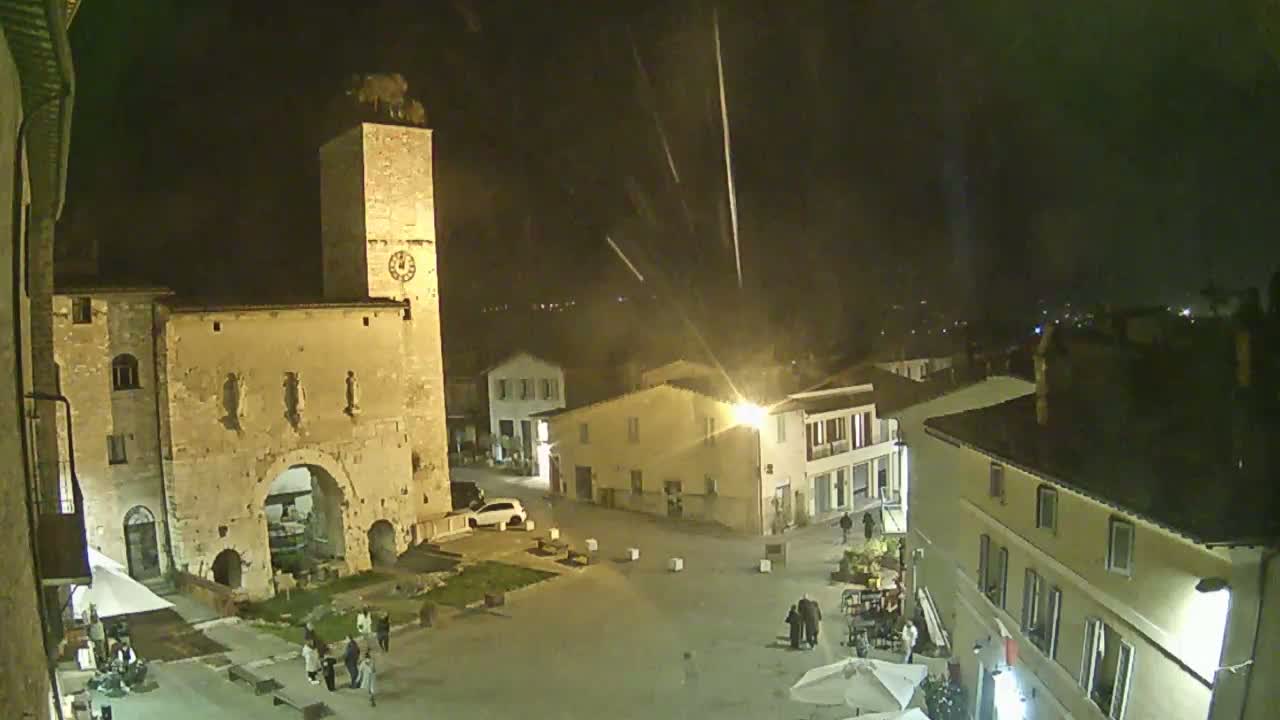 A nighttime view of a town square with a stone tower and several buildings, under light rain or snow.