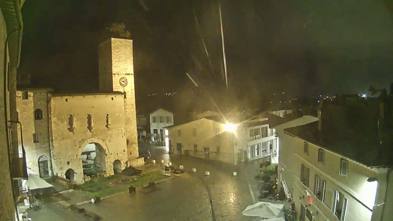 A rain-soaked town square at night, illuminated by streetlights, features an ancient stone archway and a tall clock tower.