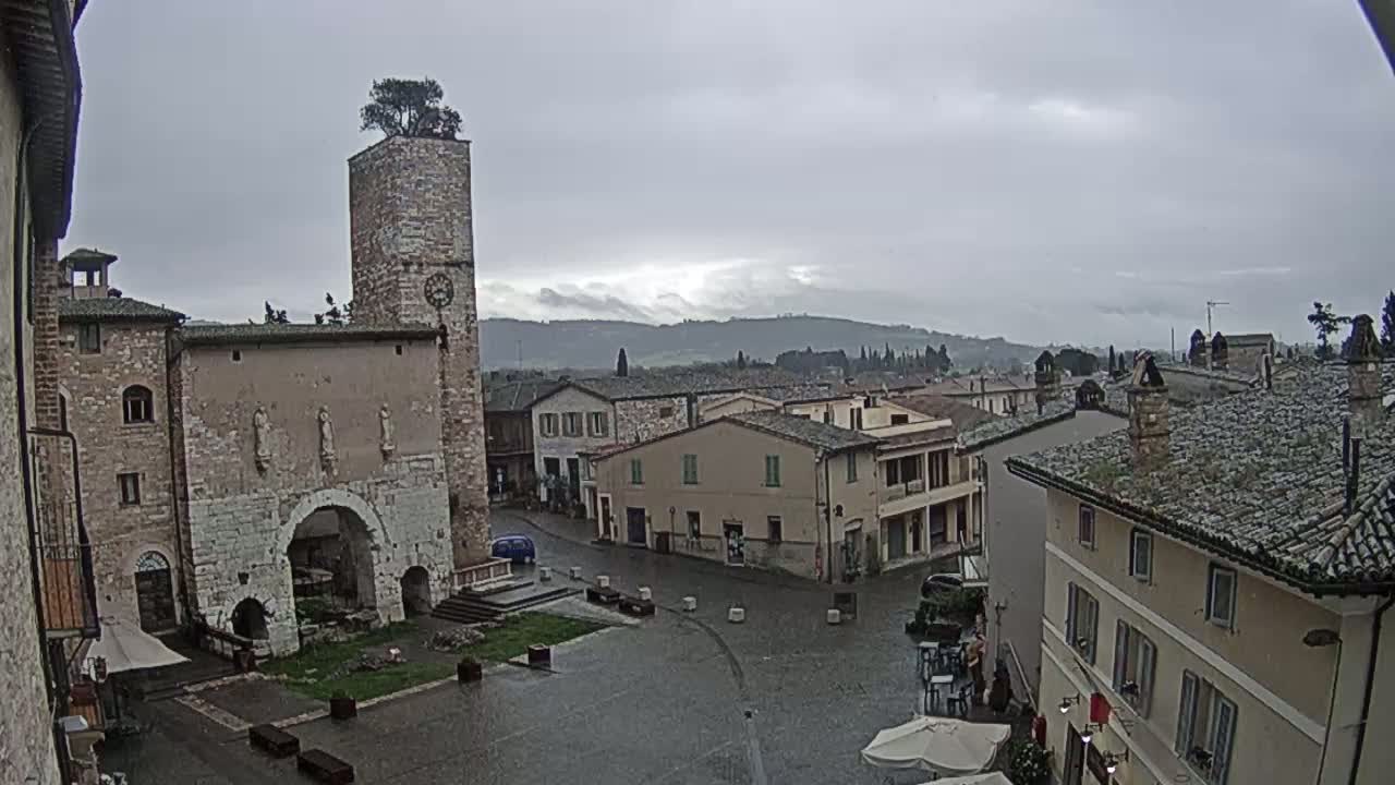 A rain-slicked town square in a historic Italian village, surrounded by stone buildings and a tall tower with a tree on top, overlooks rolling hills under a cloudy sky.