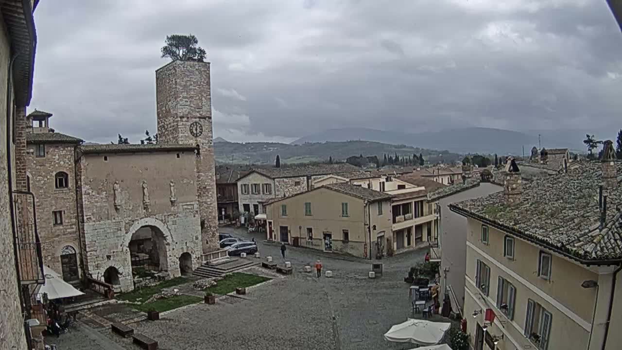 A town square in a small Italian town under an overcast sky, centered on a stone building with a clock tower topped by a tree.