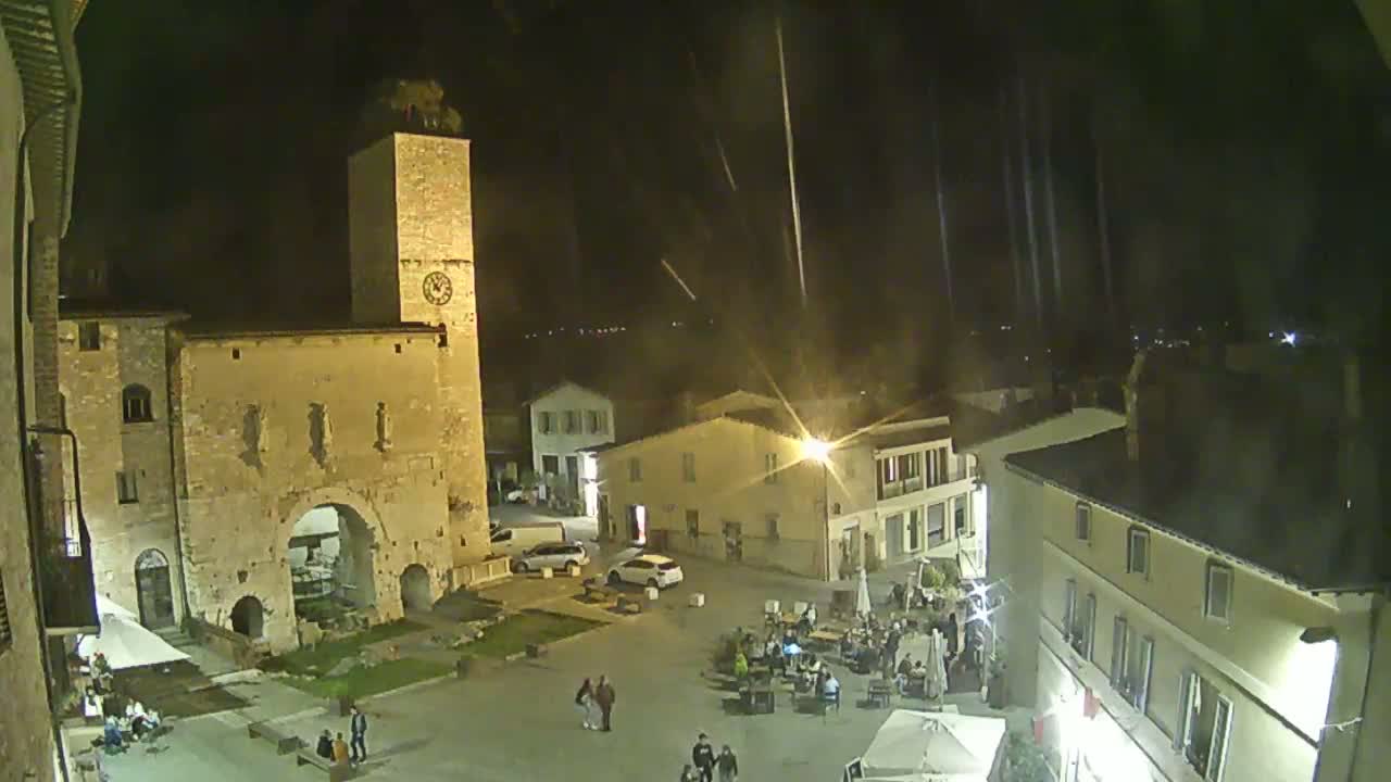 A nighttime view of a town square with several buildings, including a tall tower, and people gathered in the square under a lightly raining sky.