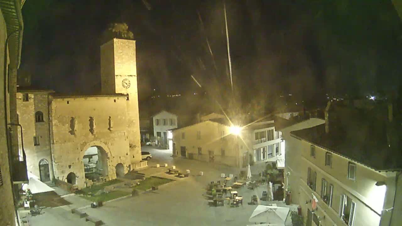 A nighttime view of a town square, illuminated by streetlights, features a tall clock tower and several buildings, with some outdoor seating visible under a possibly slightly hazy sky.