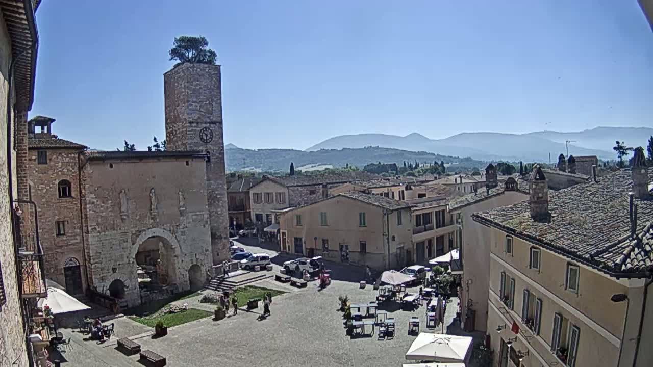 A town square with several buildings, including a tall clock tower with a tree on top, is seen under a clear, sunny sky with mountains in the distance.
