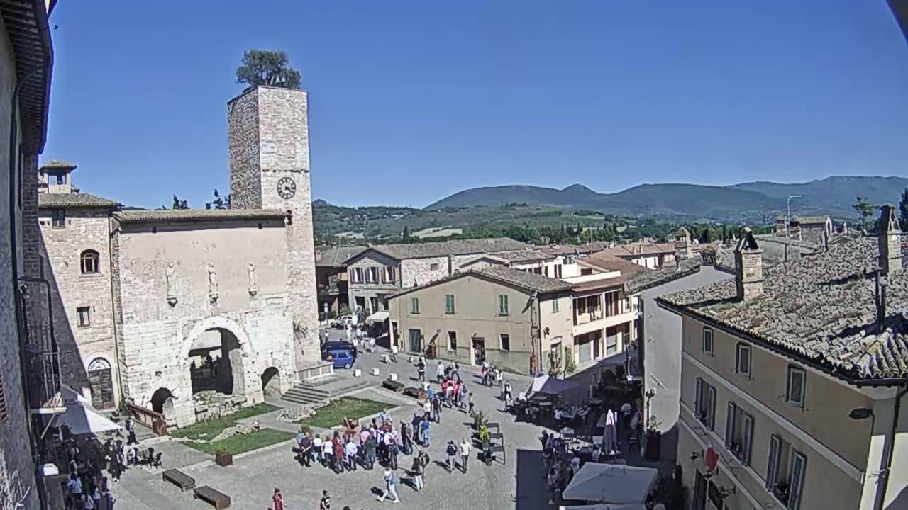 A town square with a tall clock tower, surrounded by stone buildings, under a clear blue sky, and hills visible in the distance.
