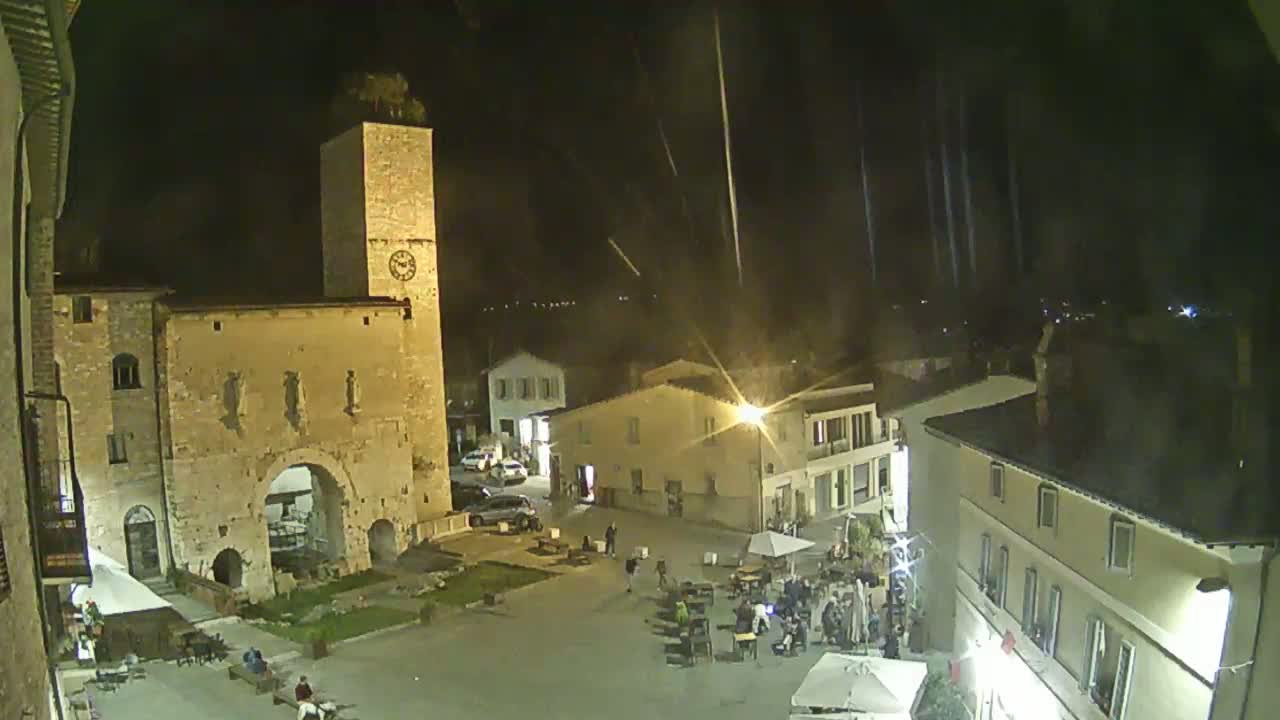 A nighttime view of a town square with several buildings, including a tall clock tower and arched entryway, under a lightly raining sky.
