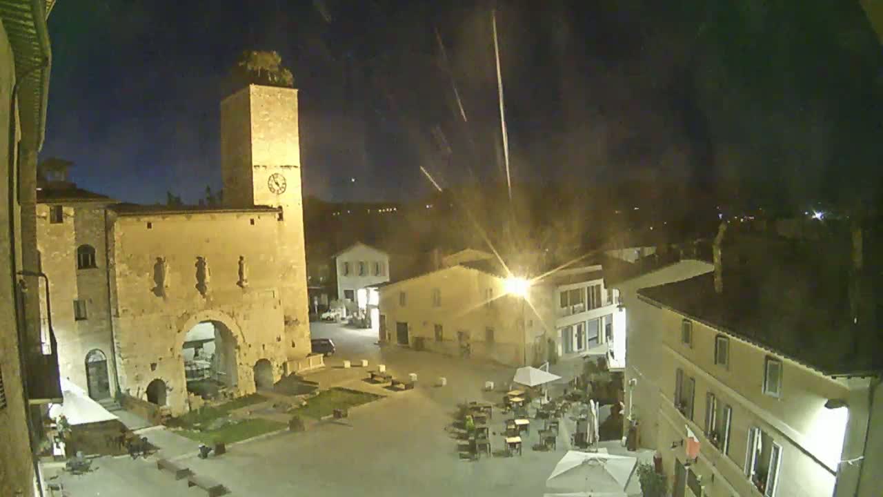 An illuminated town square at night, featuring a tall stone tower and several buildings, under a dark sky with streaks of light.