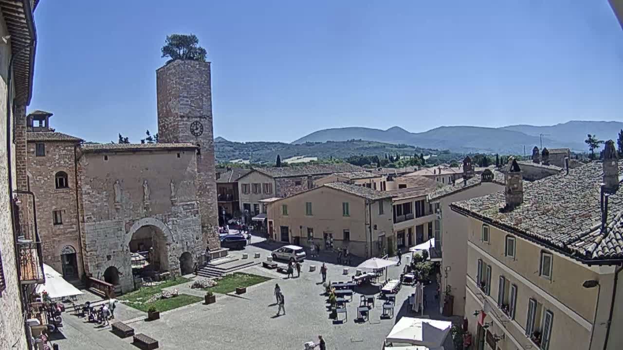 A sunny view of a town square, surrounded by stone buildings, with a tall tower topped with a tree in the background are mountains under a clear blue sky.