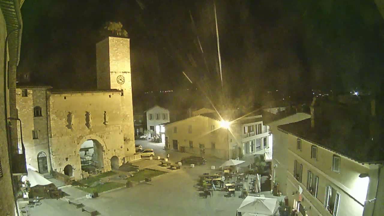 A nighttime view of a town square with a tall clock tower, buildings lit by streetlights, and several people seated at outdoor tables.