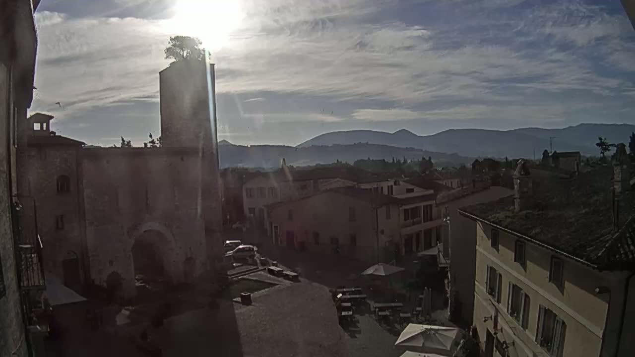 A sunlit town square, overlooked by a stone tower with a tree on top, is situated before a range of distant hills under a partly cloudy sky.