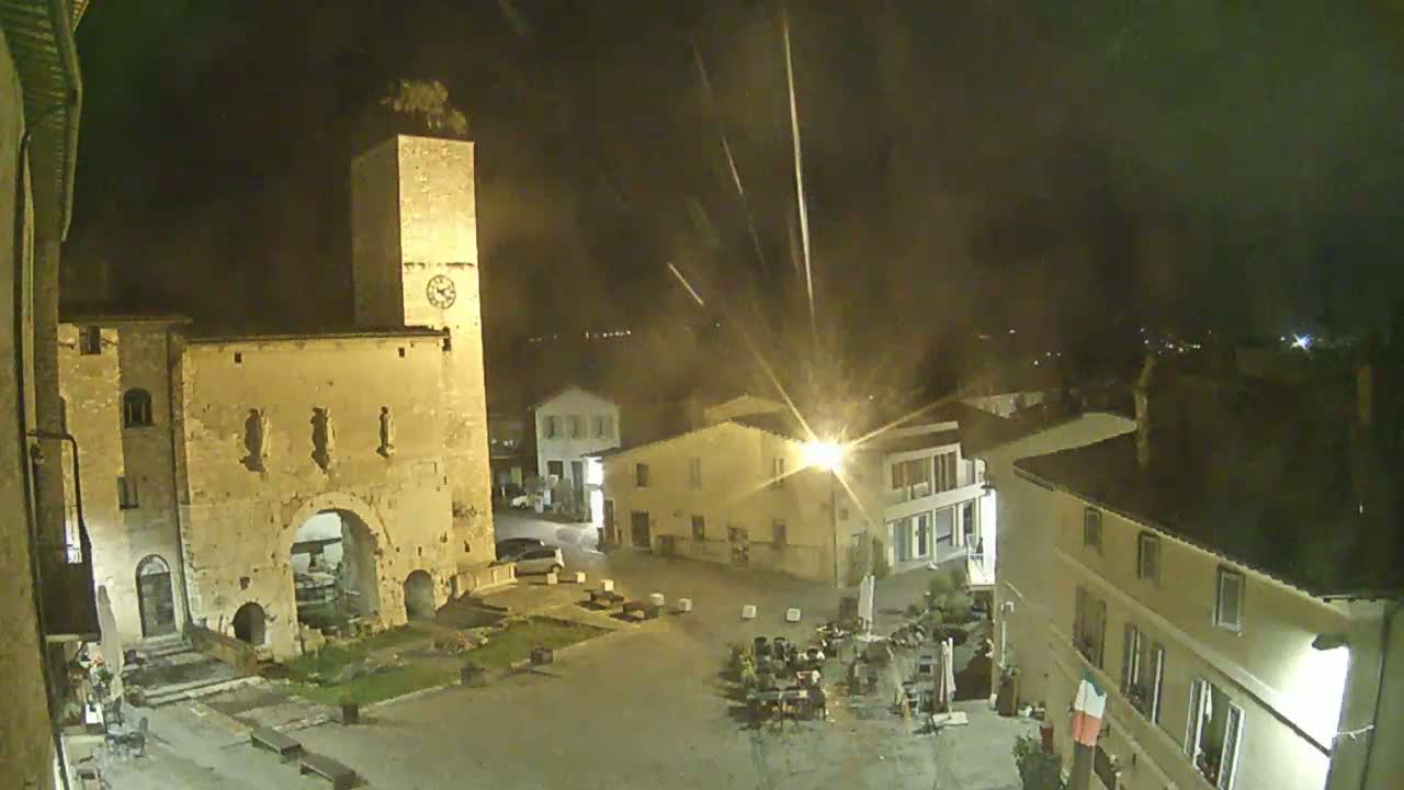 A nighttime view of a town square, featuring an ancient stone gateway and clock tower, under a slightly hazy sky.