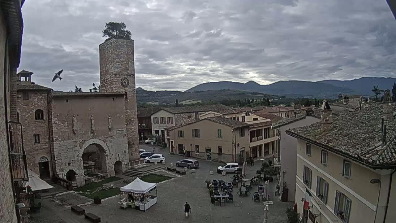 A town square in a small, stone-built town under a cloudy sky, features a tall tower, several buildings, and a few cars and people.