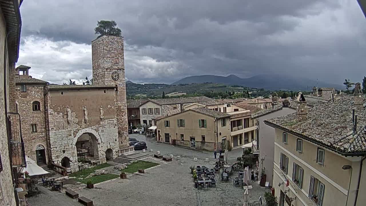 A town square in a hilly region under a cloudy sky, with stone buildings surrounding the square and a clock tower topped with a tree.