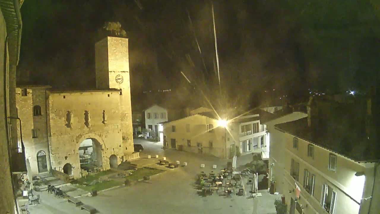 A nighttime view of a town square with a tall clock tower and several buildings illuminated under a dark, possibly slightly rainy sky.