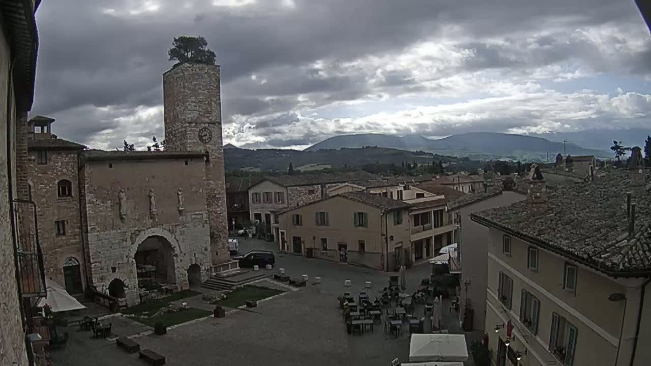 A town square with stone buildings, a clock tower topped with a tree, and outdoor seating under a cloudy sky.