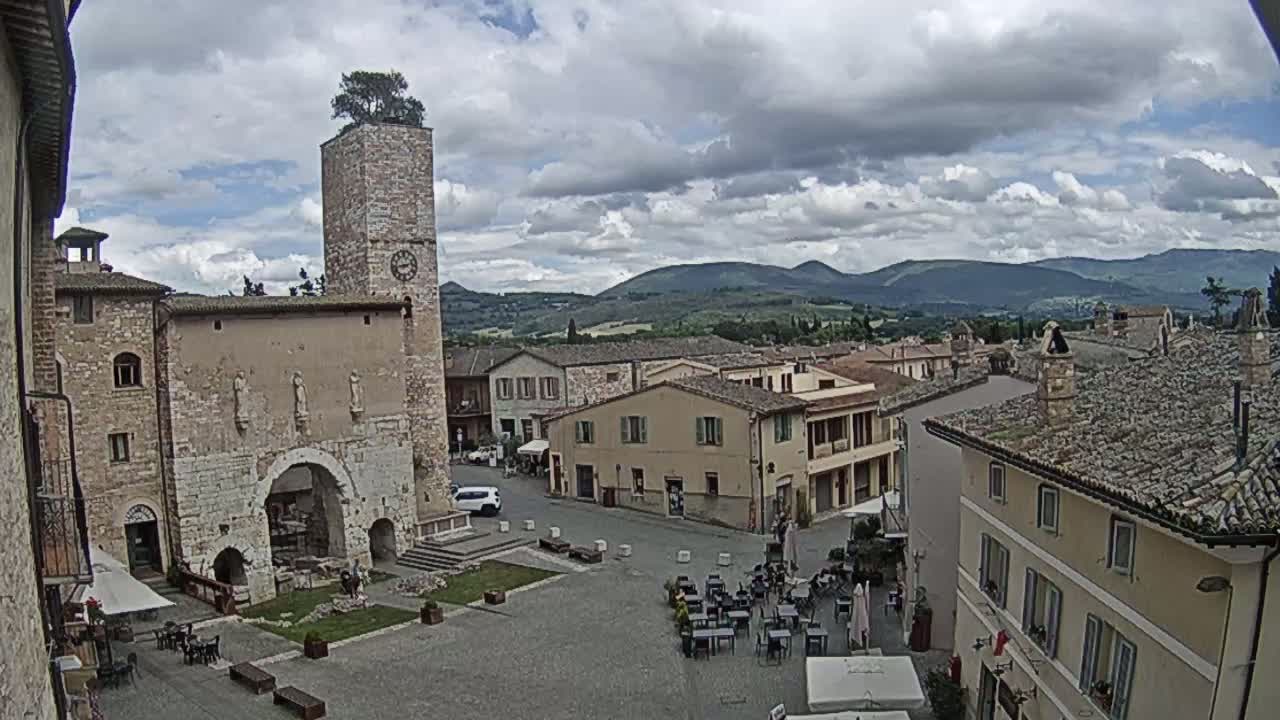 A town square in a historic Italian village, under a partly cloudy sky, is surrounded by stone buildings, one with a tall clock tower, and outdoor seating areas for cafes.