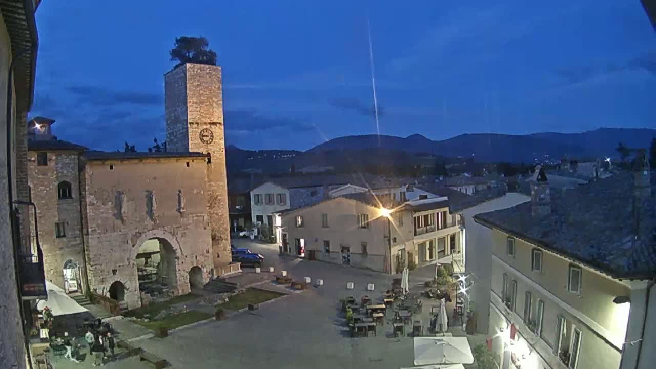 A twilight view of a town square in a small Italian village, featuring stone buildings, a clock tower with a tree on top, and outdoor seating.