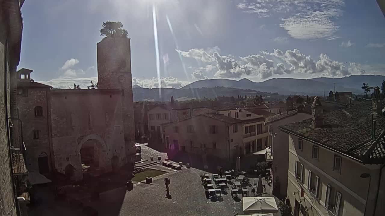 A sunny day reveals a town square in a historic Italian village, surrounded by stone buildings, with a distant mountain range visible under partly cloudy skies.
