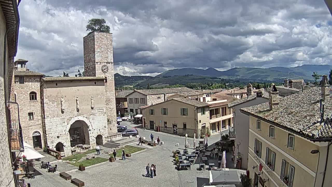 A town square in a hilly region under a partly cloudy sky, features a stone building with a clock tower and a tree on top, surrounded by other buildings and outdoor seating areas.