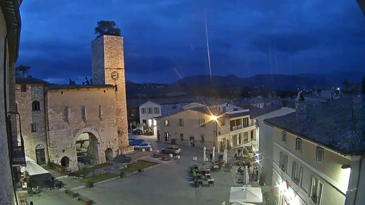 An outdoor town square at twilight is shown, with a stone clock tower and several buildings surrounding it, under a dark blue sky with some clouds.
