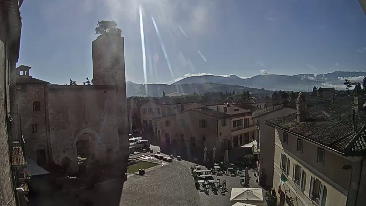 A sunny day reveals a town square surrounded by historic stone buildings, a tall tower topped with a tree, and distant mountains under a clear sky.