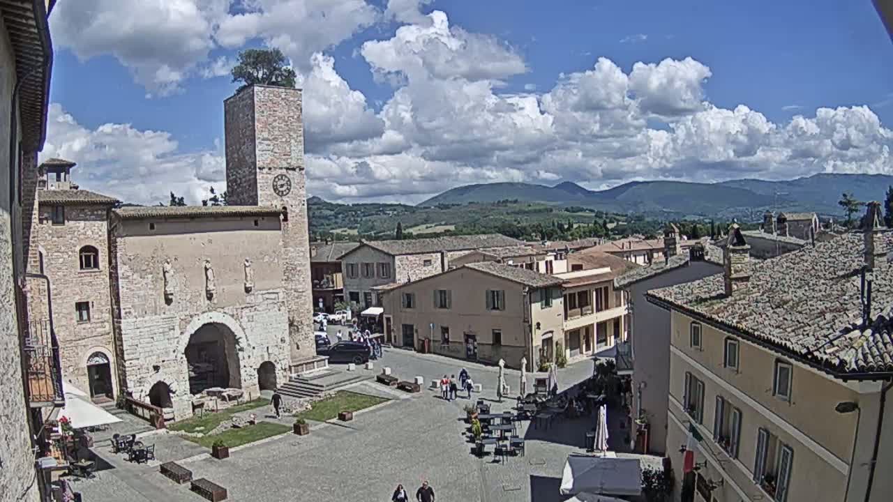 A town square in a historic village, under a partly cloudy sky, is surrounded by old stone buildings, one with a tall clock tower, and overlooks rolling hills in the distance.