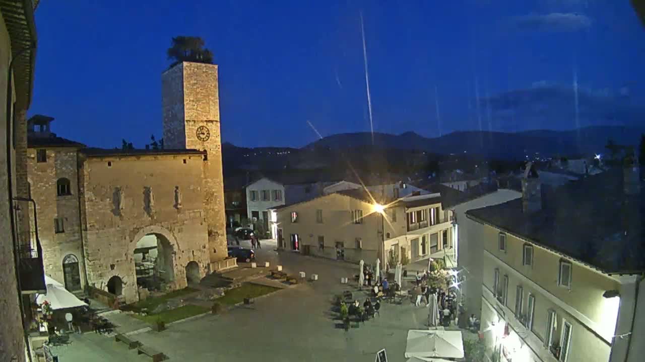 A town square at twilight, with stone buildings, a clock tower, and people sitting at outdoor tables under a clear, dark blue sky.