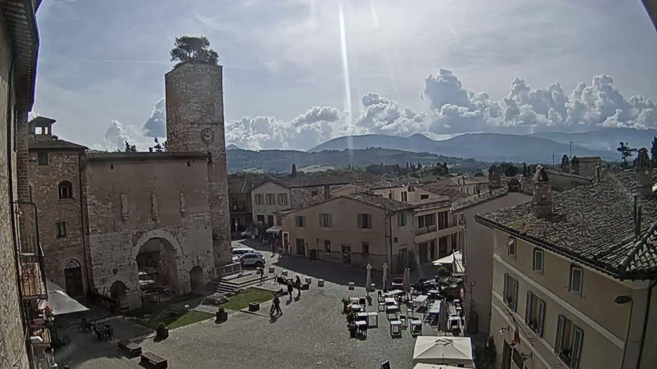A sun-drenched town square, surrounded by historic stone buildings, sits under a partly cloudy sky with distant mountains visible in the background.