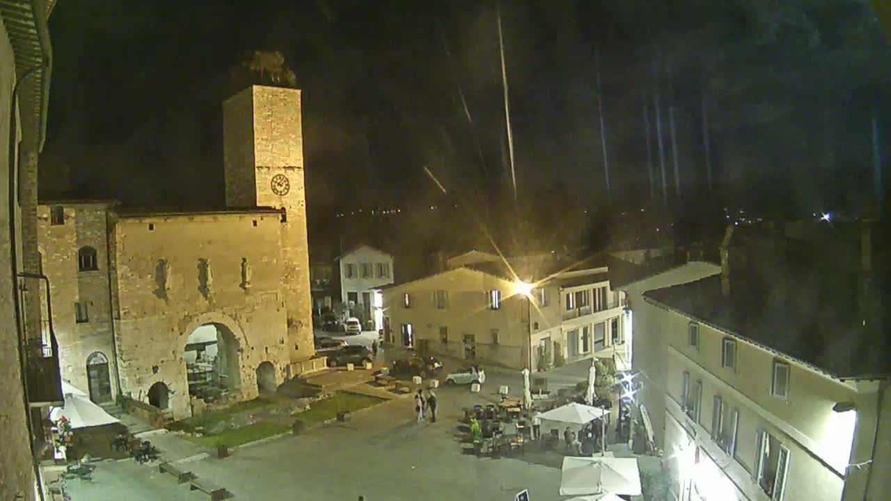 A nighttime, possibly rainy, view of a town square with several buildings, including a tall clock tower, and people sitting at outdoor tables.