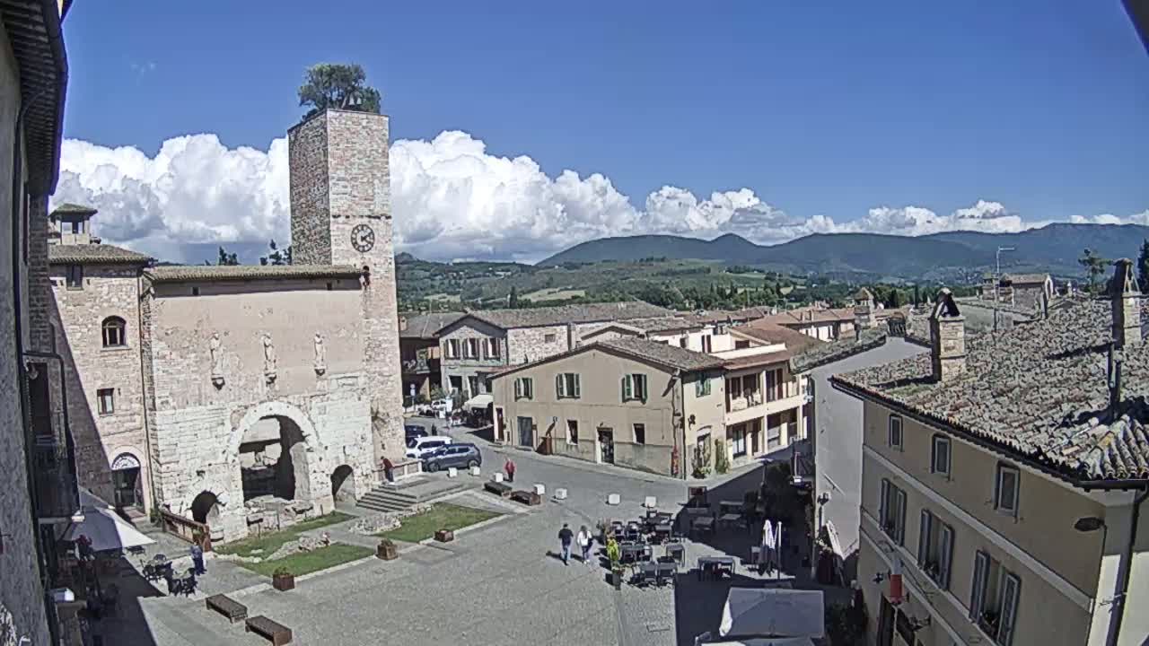 A town square with a stone tower and clock, surrounded by buildings under a partly cloudy blue sky.