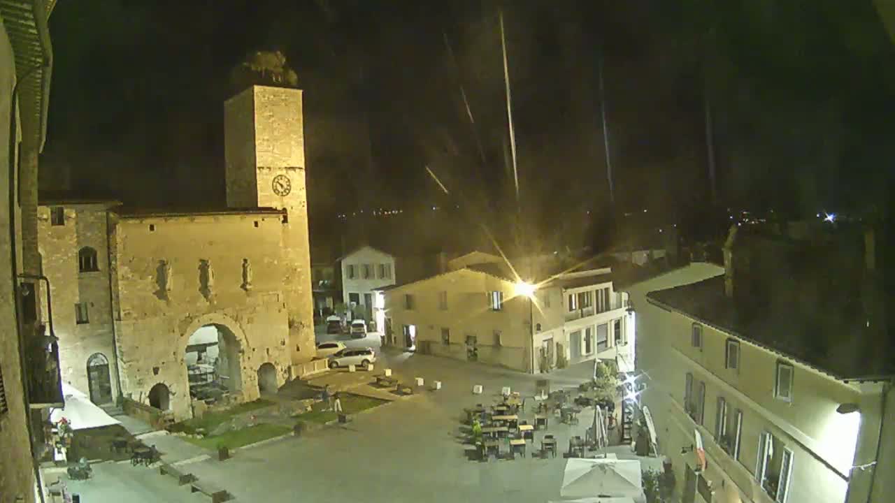 A nighttime view of a town square with several buildings, including a tall clock tower and a stone archway, under a dark sky with streaks of light.