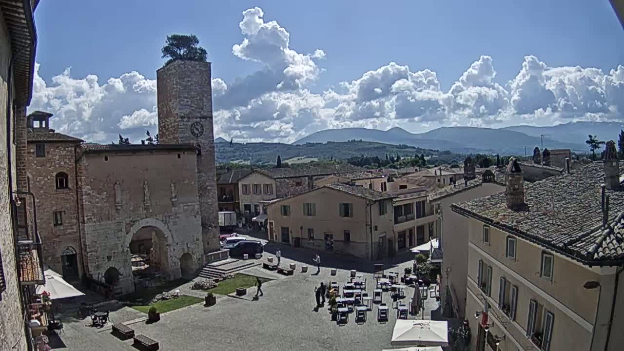 A partly sunny day reveals a town square in a historic Italian village, surrounded by stone buildings, with a distant mountain range visible under a partly cloudy sky.
