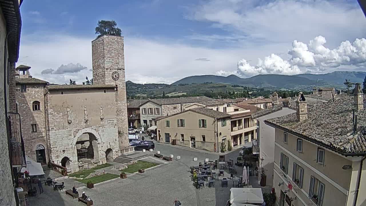 A town square in a hilly region, under partly cloudy skies, features a stone clock tower with a tree on top, surrounded by historic buildings and outdoor seating.