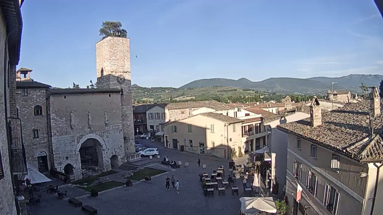 A sunny view of a town square in a historic Italian town, featuring stone buildings, a clock tower with a tree on top, and people sitting at outdoor cafes.