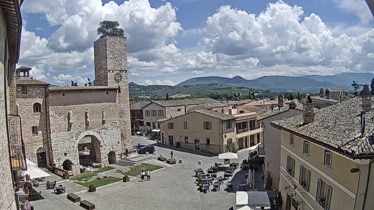 A town square with a stone tower and surrounding buildings, under a partly cloudy sky, overlooks a hilly landscape in the distance.