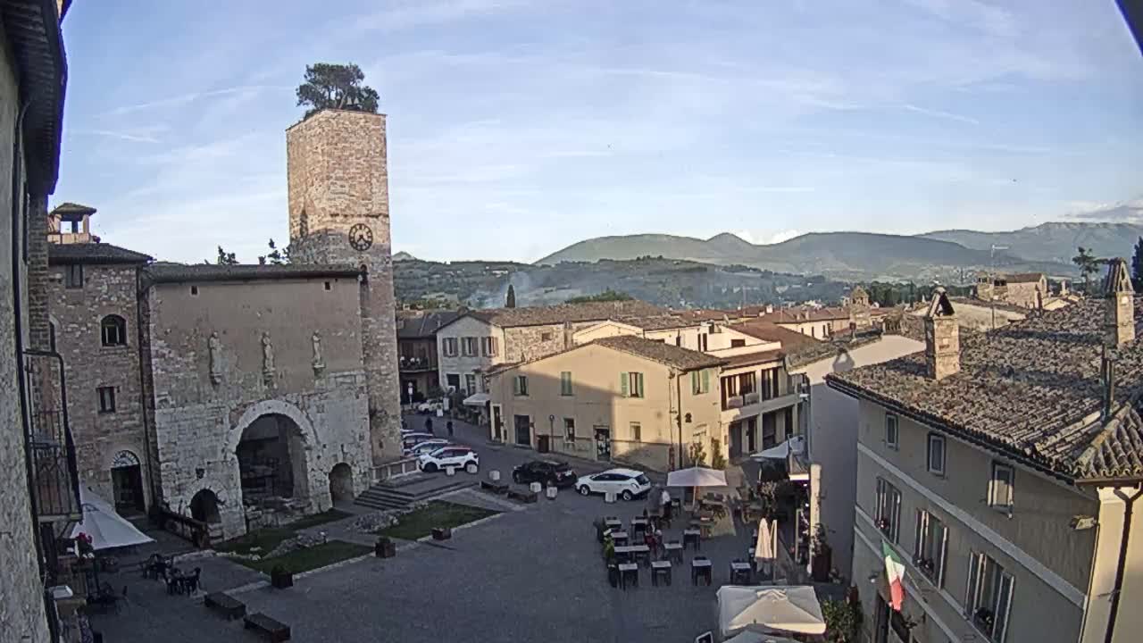 A town square with stone buildings, a clock tower topped with a tree, and several cars is visible, with distant mountains under a clear, sunny sky.