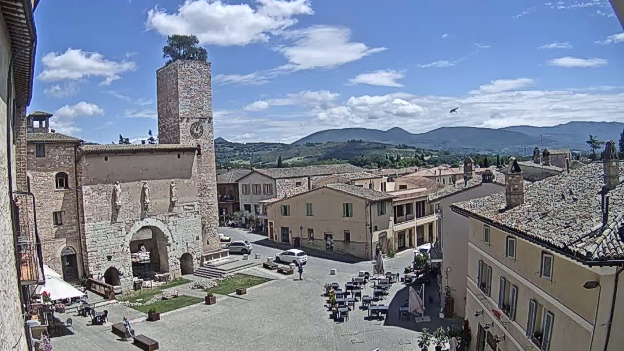 A town square with stone buildings, a clock tower topped with a tree, and outdoor seating under a partly cloudy, sunny sky, with mountains in the distance.