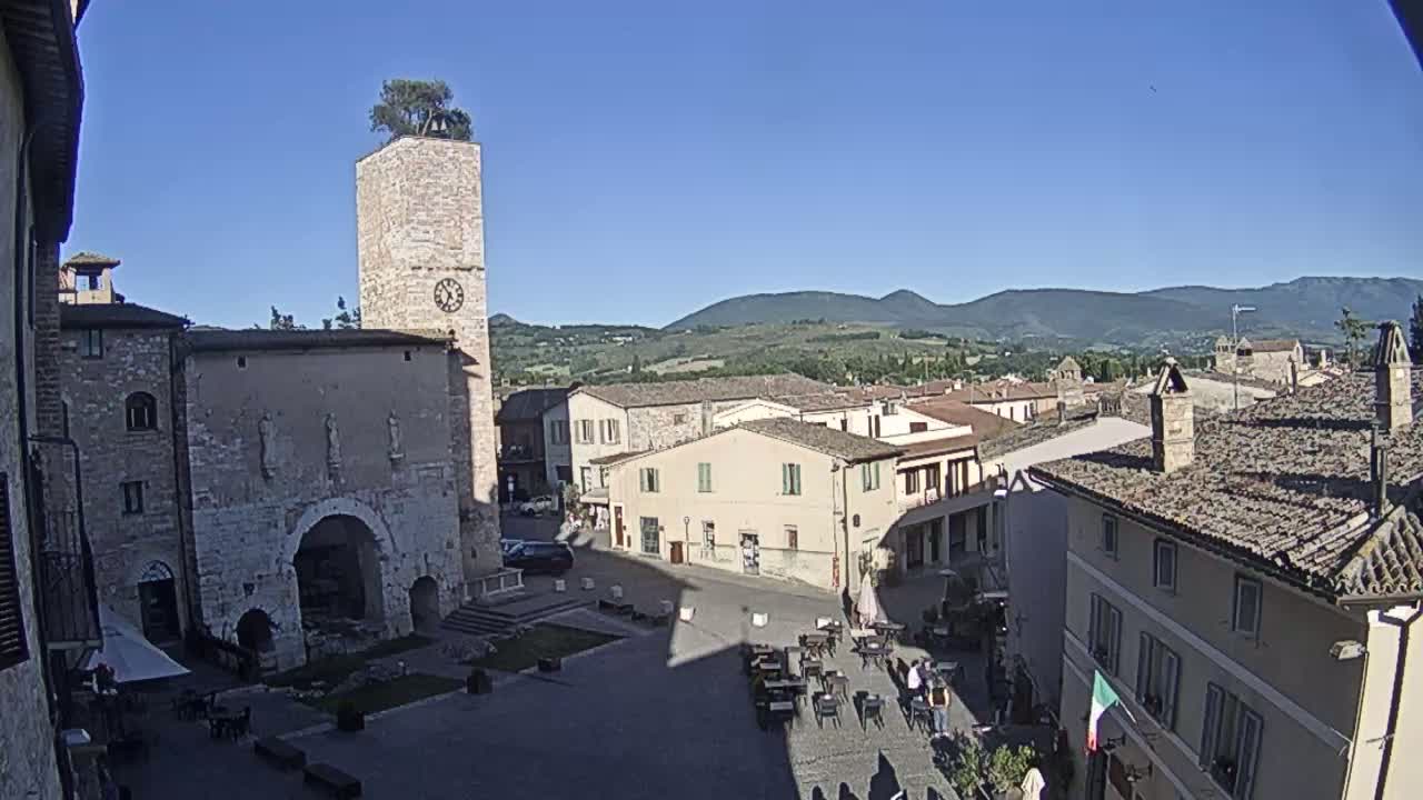 A town square with stone buildings, a clock tower topped with a tree, and outdoor seating under a clear blue sky.
