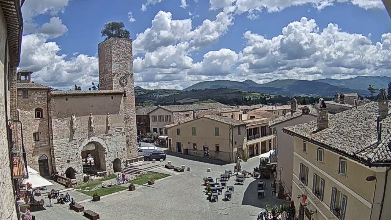 A town square in a small Italian village, under a partly cloudy sky, is surrounded by stone buildings, with a distant view of rolling hills.